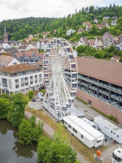 The Ferris wheel stands prominently in an urban landscape with a river and wooded hills, Ferris