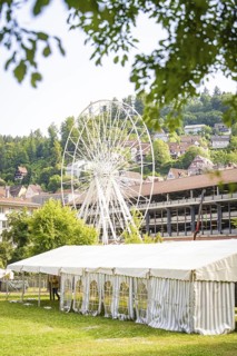 Ferris wheel behind a large white tent, surrounded by village houses and green hills, Ferris wheel