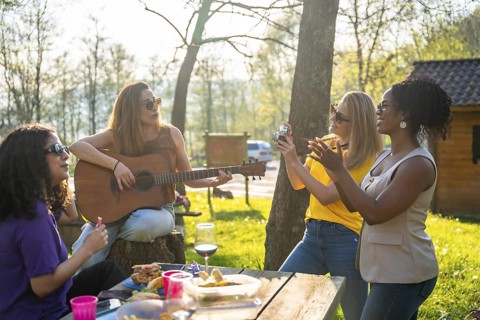 Four friends are having a picnic outdoors, with one playing guitar, one taking photos, and the