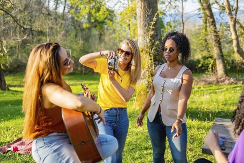 Group of friends soaking up the sun in nature, playing music, capturing moments with cameras, and