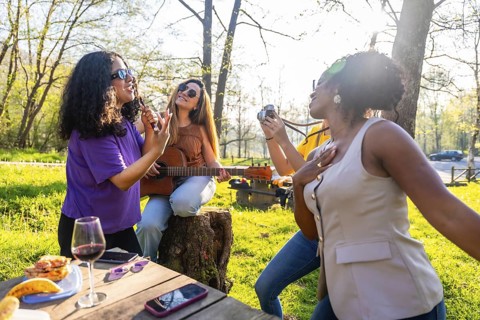 Four female friends enjoying a picnic in nature, singing, playing guitar and taking photos