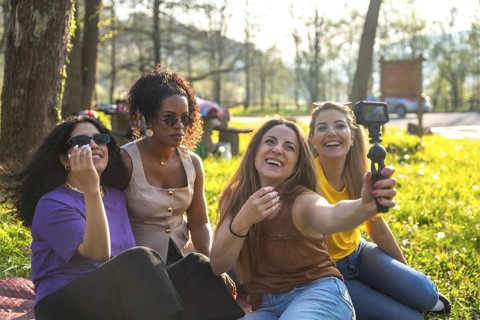 Four multi ethnic female friends are sitting on grass in a park, taking a selfie with an action