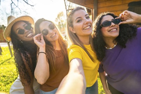 Four friends are taking a selfie outdoors, wearing sunglasses and smiling, enjoying a sunny day and