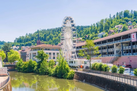 A white Ferris wheel against an urban backdrop and a green, wooded hill in bright sunlight, 950