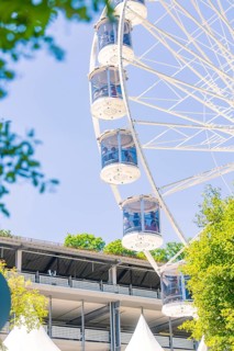 The Ferris wheel with glass cabins rises next to a large building surrounded by sunny trees, 950
