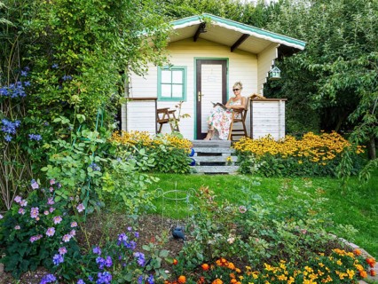 A woman in a dress sits relaxed on the small veranda of a garden shed and reads. Blooming summer