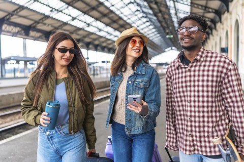 Three young tourists are walking through a train station, pulling suitcases and carrying a