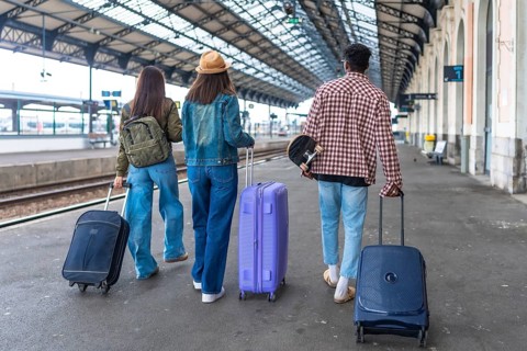 Three young tourists are walking away from camera in a train station with their suitcases and a