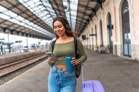 Young woman tourist walking in train station using smartphone and headphones while carrying