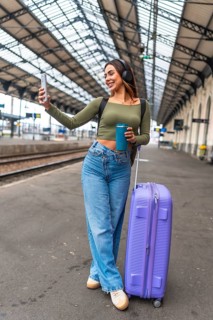 Tourist taking selfie with smartphone while waiting for train arrival at station platform, holding