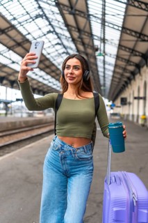 Tourist taking selfie with smartphone while walking in train station with luggage and reusable