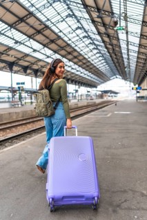 Tourist wearing headphones walking in train station pulling a purple suitcase and carrying a