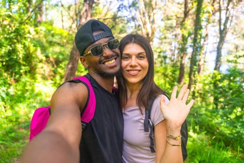 Interracial couple smiling and waving while taking a selfie during a vibrant hike in a lush, green