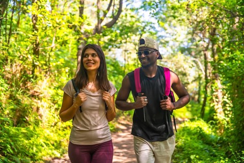 Interracial couple wearing backpacks walking in a forest trail, enjoying the beautiful nature