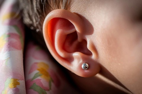 Delicate silver earring stud in baby's ear bathed in warm golden light. Tender moment with subtle