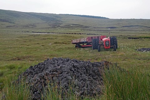 Old red tractor standing in boggy fields against a hilly backdrop with grey sky, peat cutting, Isle