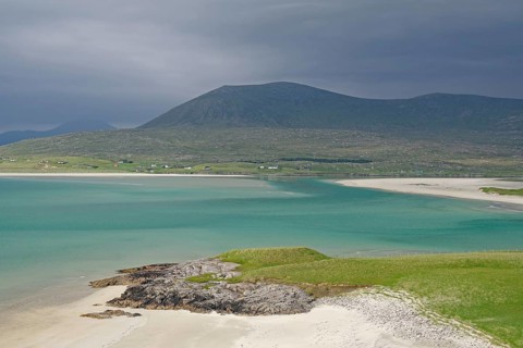 Wide sandy beach meets turquoise waters, surrounded by green hills and mountains under a gloomy
