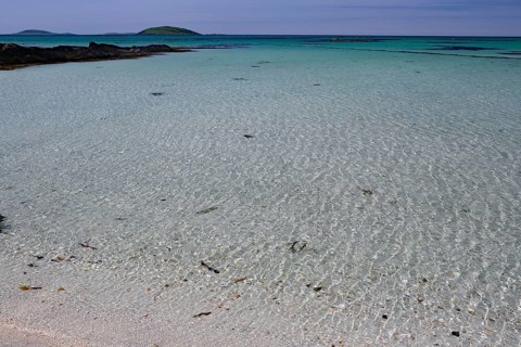 Crystal clear turquoise water on a sandy beach with islands on the horizon under a blue sky,