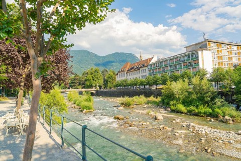 A calm river flows along an urban promenade with mountains in the background, Merano, Italy