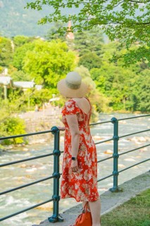 A woman in a flowered dress looks at a flowing river on a sunny day, Merano, Italy