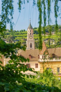 Church tower and building surrounded by green trees and hills, Merano, Italy