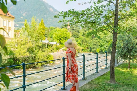 A woman looks at a river in the middle of a green, summery landscape, Merano, Italy