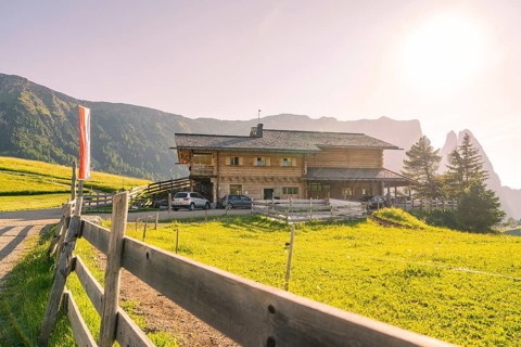 Large farmhouse with wooden fence on a wide green field in a sunny mountain landscape, Alpe di