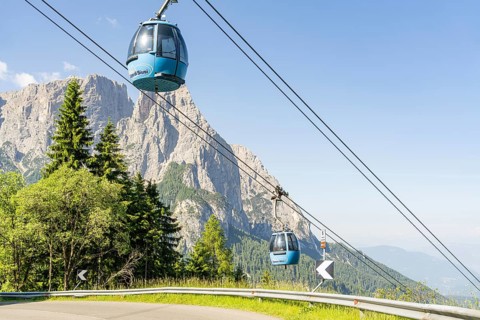 Two blue cable car gondolas hovering over trees in front of a rocky mountain range, Alpe di Siusi,