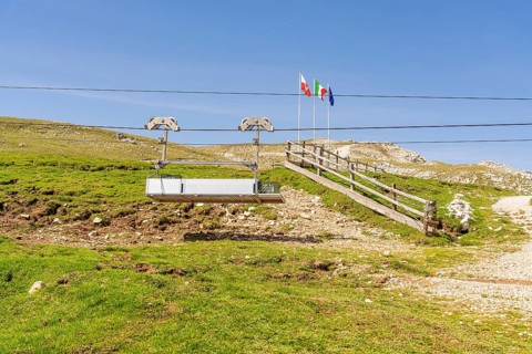 Chairlift in front of mountain landscape with flags and green slope under clear blue sky, Alpe di