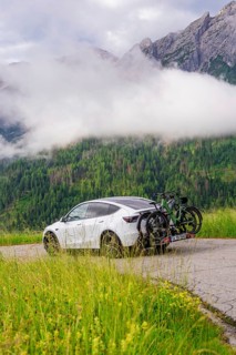 White car with bicycles on road, surrounded by misty mountain landscape and green forest, electric