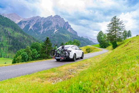 White car with bicycles on a road through green, hilly landscape with cloudy sky, electric car on