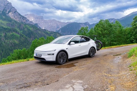 White car with bicycles on a winding mountain road, surrounded by dense greenery and cloudy sky,