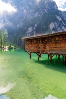 A wooden house stands on stilts above a clear, green mountain lake, surrounded by tall trees and