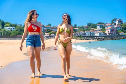 Two cheerful young women wearing bikinis and sunglasses are enjoying a walk on a sunny beach,
