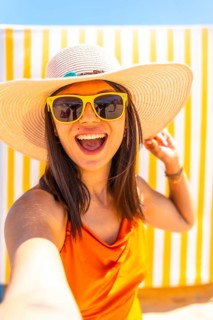Young woman wearing sunglasses and a straw hat taking a selfie while smiling at the camera on a