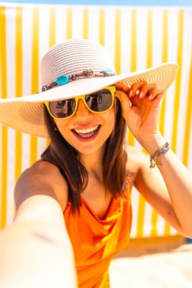 Young woman taking a selfie on a sunny beach, wearing an orange dress and a wide brimmed sun hat