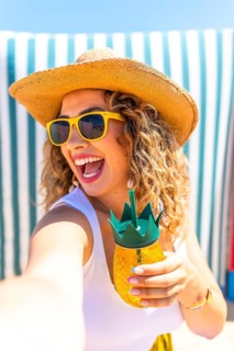 Happy tourist woman wearing sunglasses and straw hat taking a selfie at beach while holding a