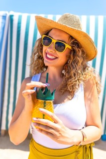 Young curly blonde woman wearing sunglasses and straw hat drinking juice at beach bar, enjoying