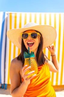 Young woman enjoying a refreshing pineapple cocktail while relaxing on the beach, wearing stylish