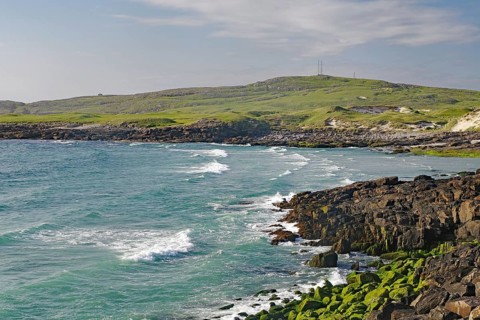 Rocky coastline with strong waves and green grassland under a cloudy sky, wild, Isle of Barra,