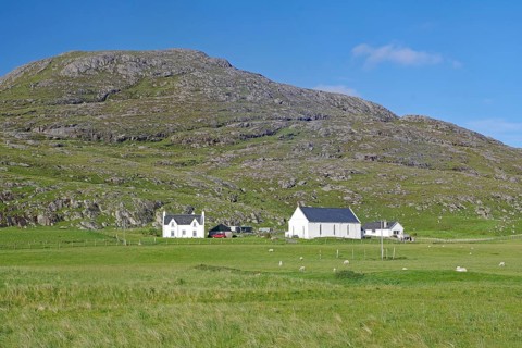 White houses stand in a green meadow with mountains in the background. Sheep grazing nearby, Barra,