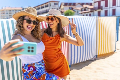 Two cheerful women wearing summer hats and sunglasses are taking a selfie with a smartphone on a