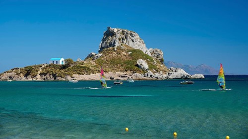 Island with windsurfers and boats in the turquoise blue sea in front of a rocky coast, Agios