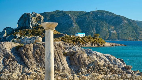 Ancient column and stones in front of island with chapel, blue sky in the background, Agios