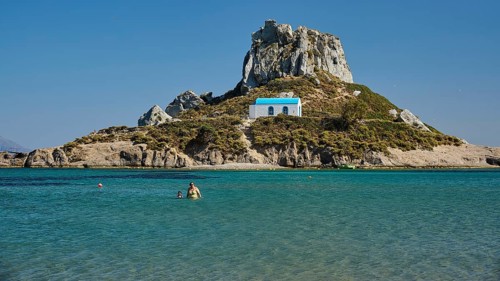 Small chapel with blue roof on rocky island in clear sea, Agios Stefanos Beach, Agios Stefanos