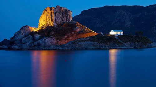 An idyllic night view of an illuminated island with a chapel and calm sea, Agios Stefanos Beach,