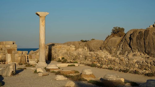 Ancient ruins with a standing stone column and surrounding rock structures in warm sunlight, Agios