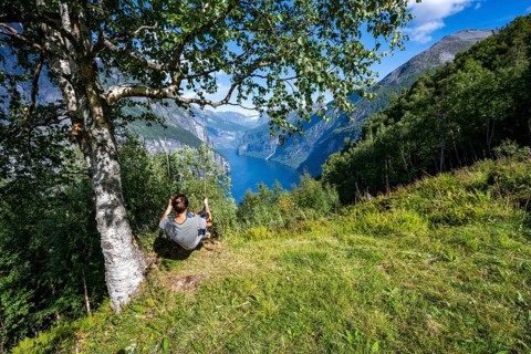 Young man swinging on a tree, view of idyllic fjord landscape of the Geirangerfjord at Blomberg