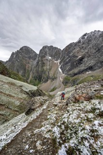 Mountaineer on hiking trail in steep rocky mountain landscape with fresh snow in summer, behind