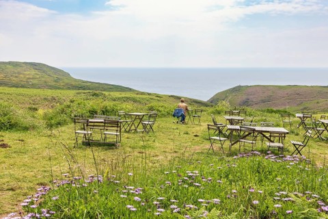Outdoor cafe with a alone woman sitting by a table on a meadow with a sea view in the summer,